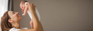 Mother holds a newborn up in the air by a window and beige wall