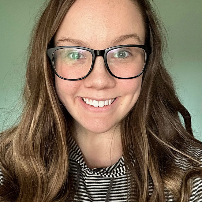 Headshot of Ryleigh Cochran from Gold Coast Doulas wearing black framed glasses and a black and white striped hoodie against a green wall