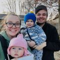 Ryleigh from Gold Coast Doulas poses with her family wearing a forward facing infant and her husband beside her holding a toddler in front of their home