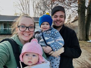 Ryleigh from Gold Coast Doulas poses with her family wearing a forward facing infant and her husband beside her holding a toddler in front of their home