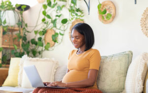 Pregnant mom wearing a yellow t-shirt sitting on a sofa, working on a laptop, with greenery against the wall behind her