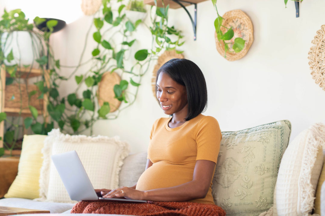 Pregnant woman wearing a yellow t-shirt sitting on a beige sofa working on a laptop with greenery on the wall behind her