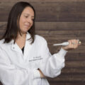 Dr. Amy holding device while wearing her white coat against a wooden wall