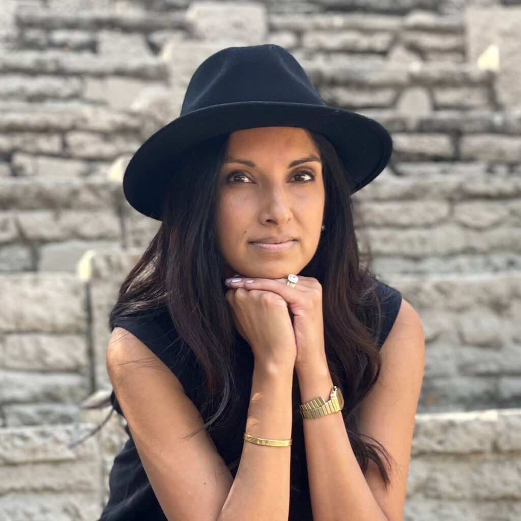 Payal Adhikari resting her head on her hands wearing a black hat and black top against a brick wall