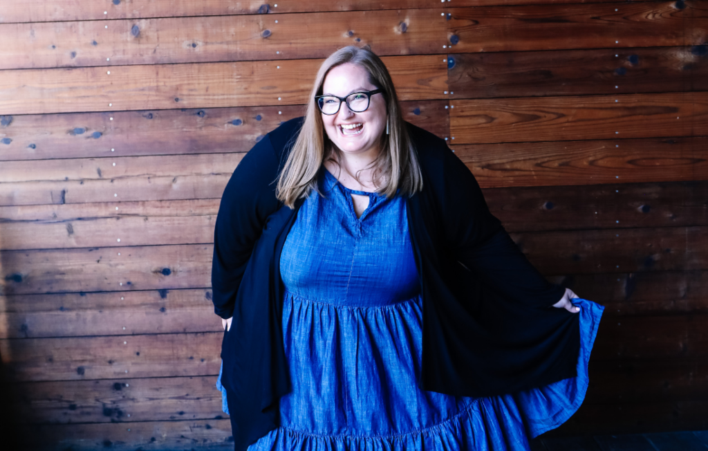 Plus size woman wearing a blue dress and black cardigan smiling against a wooden wall