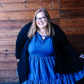 Plus size woman wearing a blue dress and black cardigan smiling against a wooden wall