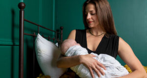 Woman holding a swaddled baby on a yellow bed with green walls in the background