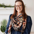 Red headed woman smiles with red glasses in a jean dress that's belted with a printed scarf in front of a white wall, fireplace, and greenery