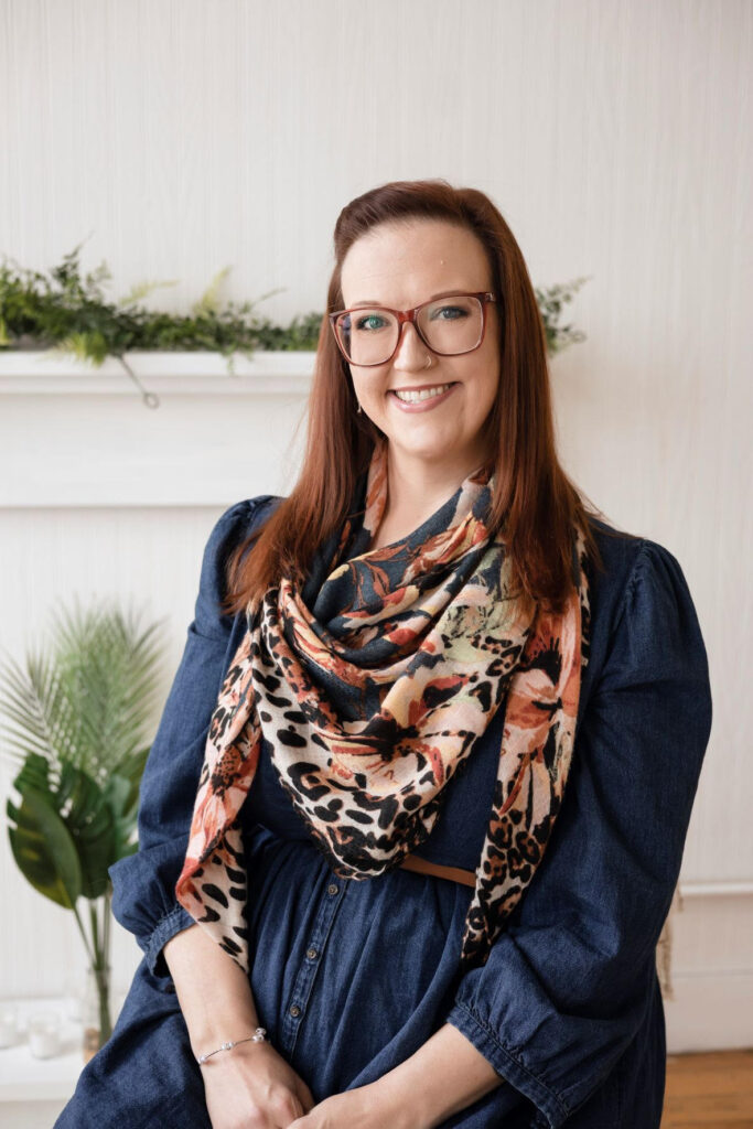 Red headed woman smiles with red glasses in a jean dress that's belted with a printed scarf in front of a white wall, fireplace, and greenery