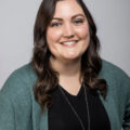 Kristin Morter from Gold Coast Doulas smiles wearing a black v-neck, long necklace, and teal cardigan against a grey background