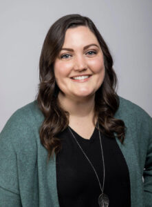 Kristin Morter from Gold Coast Doulas smiles wearing a black v-neck, long necklace, and teal cardigan against a grey background