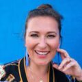 Smiling woman wearing bright blue earrings, a black top, and hand resting on her cheek with a blue wall behind her