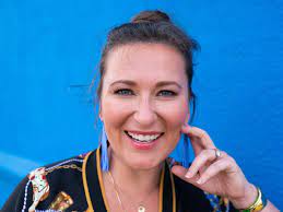 Smiling woman wearing bright blue earrings, a black top, and hand resting on her cheek with a blue wall behind her