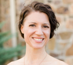 Headshot of Katie Timbrook of Athena's Bumb wearing a gold necklace with brick backround