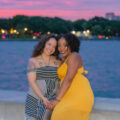 Jade Sanchez from Gold Coast Doulas wearing a yellow summer dress poses with a woman wearing a striped maxi dress at sunset by a body of water