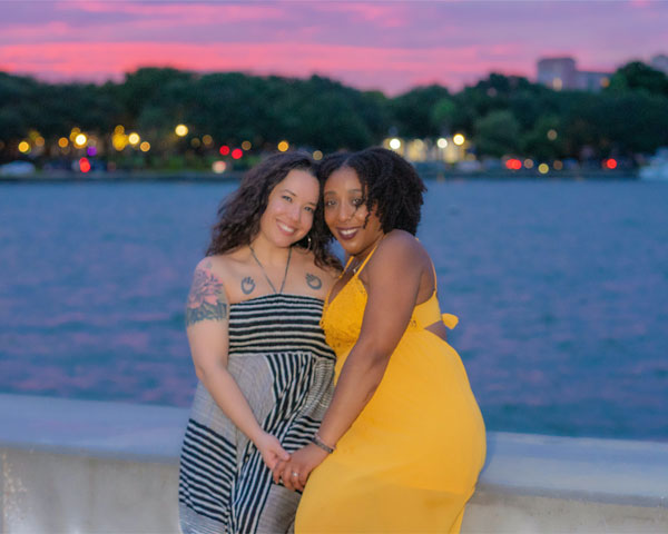 Jade Sanchez from Gold Coast Doulas wearing a yellow summer dress poses with a woman wearing a striped maxi dress at sunset by a body of water
