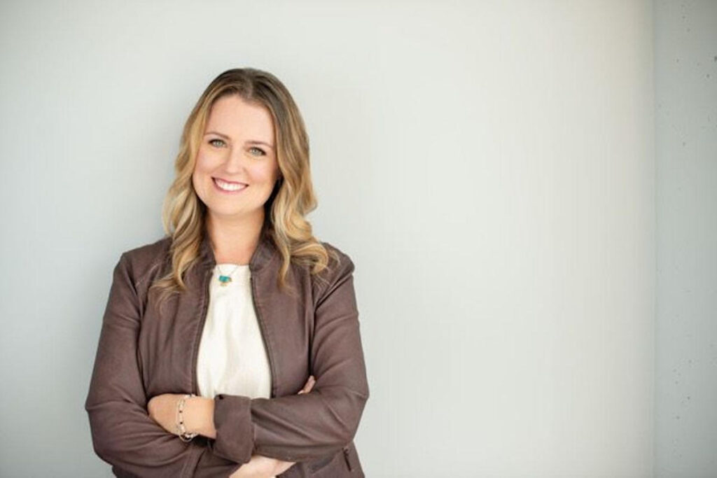 Laura Fletcher poses with her arms crossed wearing a white blouse and brown blazer against a white wall