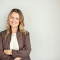 Laura Fletcher poses with her arms crossed wearing a white blouse and brown blazer against a white wall