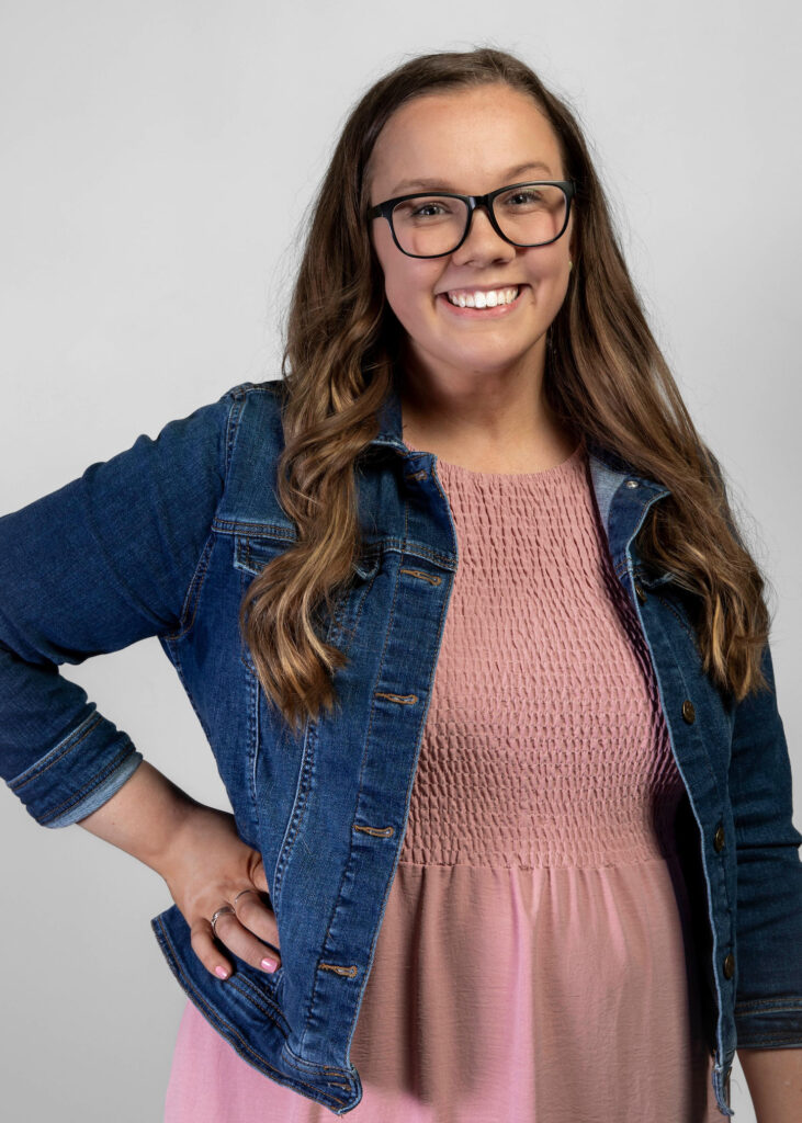 Ryleigh Cochran poses with a hand on her hip wearing a mauve dress and jean jacket against a grey background