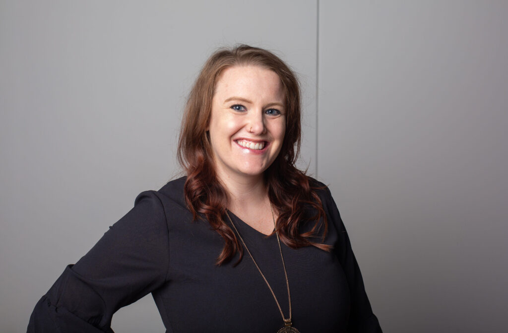 Woman wearing a long gold necklace, black blouse, and nose ring smiles in front of a light purple wall