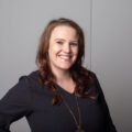 Woman wearing a long gold necklace, black blouse, and nose ring smiles in front of a light purple wall