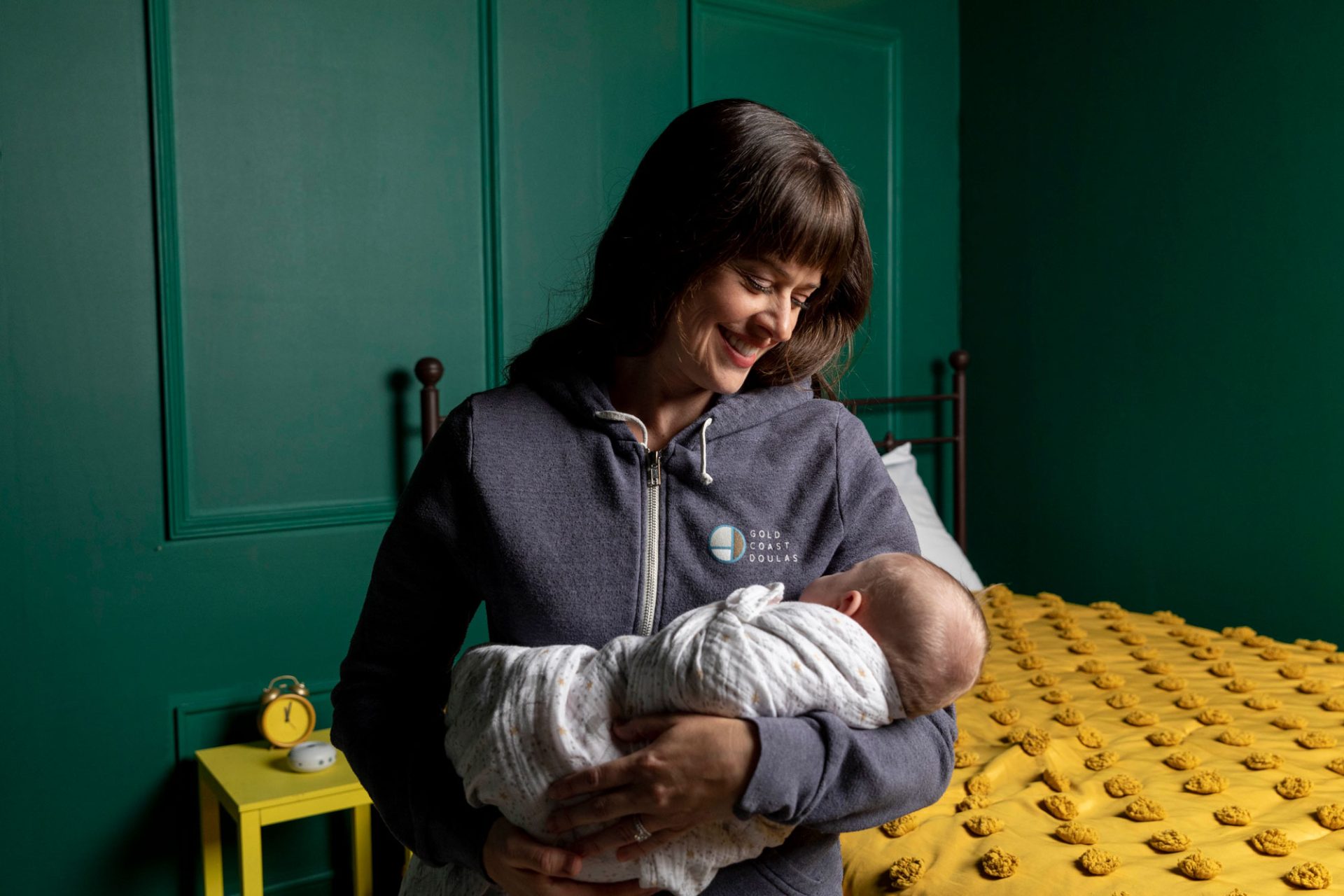 Kay from Gold Coast Doulas wearing a GCD zip up hoodie holding a sleeping baby with a yellow bed and green walls in the background