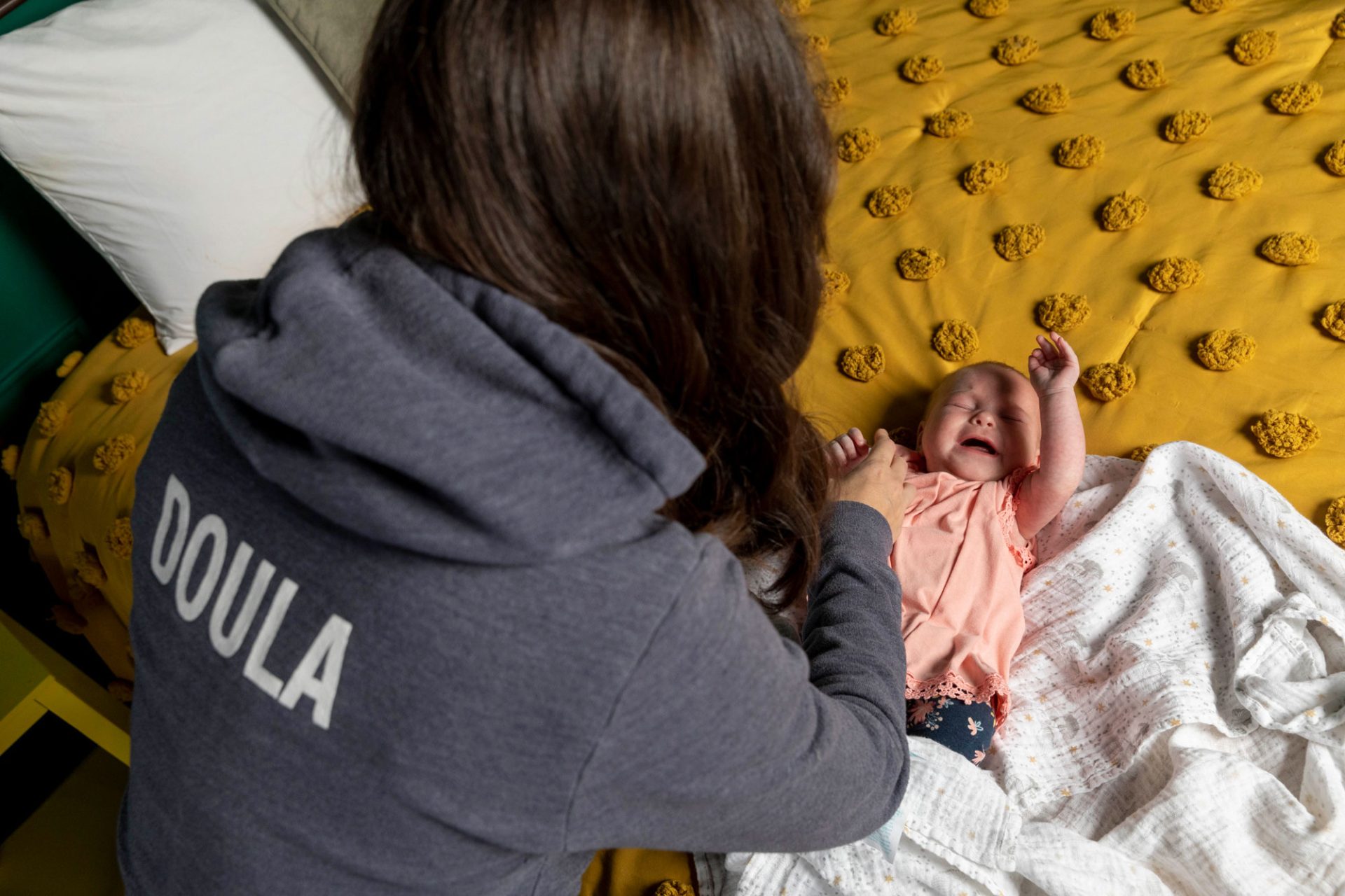 Kay from Gold Coast Doulas wearing a doula hoodie, swaddling a wiggly baby on a yellow bed