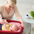 Bebcare - There Beyond Touch. Picture of a mom smiling and looking at her baby that's laying down in a bassinet and holding her hand.