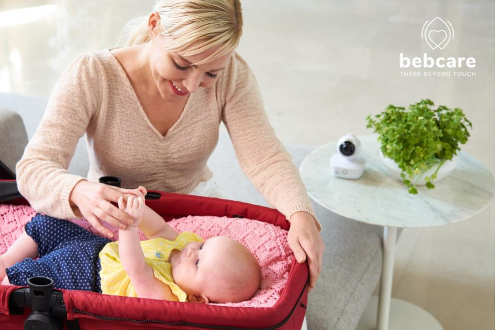 Bebcare - There Beyond Touch. Picture of a mom smiling and looking at her baby that's laying down in a bassinet and holding her hand.