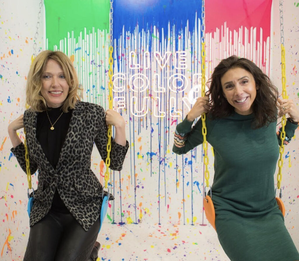 Kristin in black and Alyssa in teal sitting on swings with splatter painted background that states "Live Colorfully"