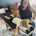 Dr. Annie Bishop wearing a black tank top and jeans giving a chiropractic adjustment to a little girl with a yellow flower, white shirt, and navy blue shorts
