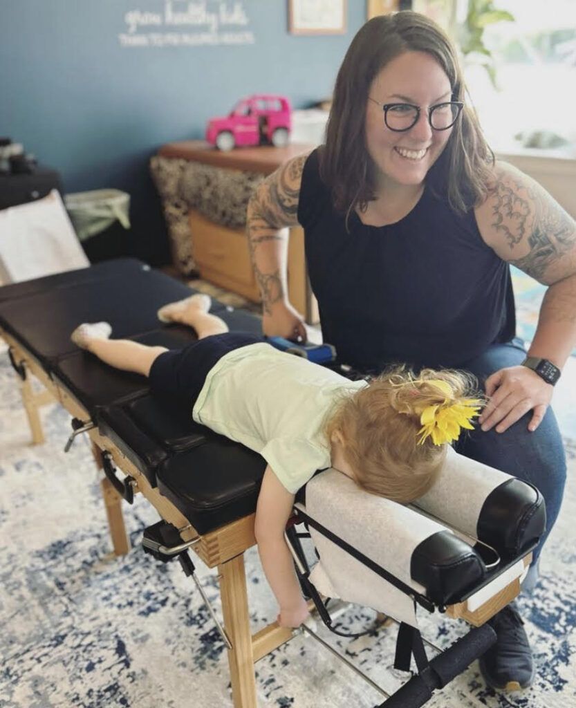 Dr. Annie Bishop wearing a black tank top and jeans giving a chiropractic adjustment to a little girl with a yellow flower, white shirt, and navy blue shorts