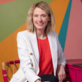 Kristin from Gold Coast Doulas wearing a red blouse and a white blazer sitting in front of a multi-colored wall