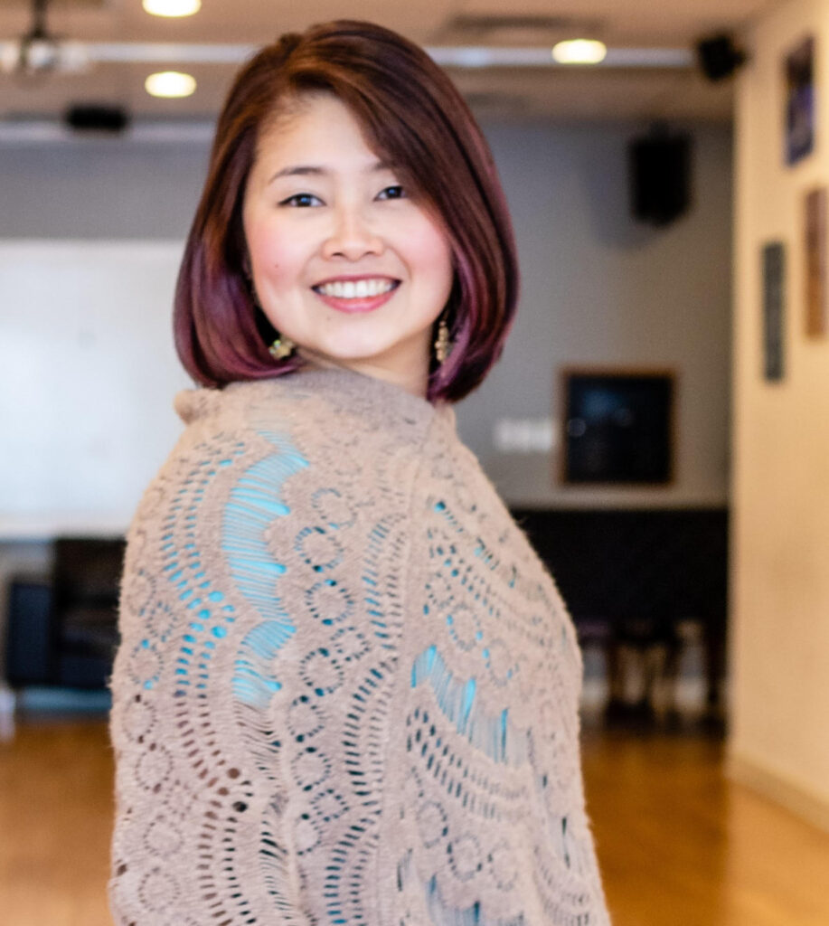 Regina Lum wearing mauve and teal clothing smiles in front of a studio