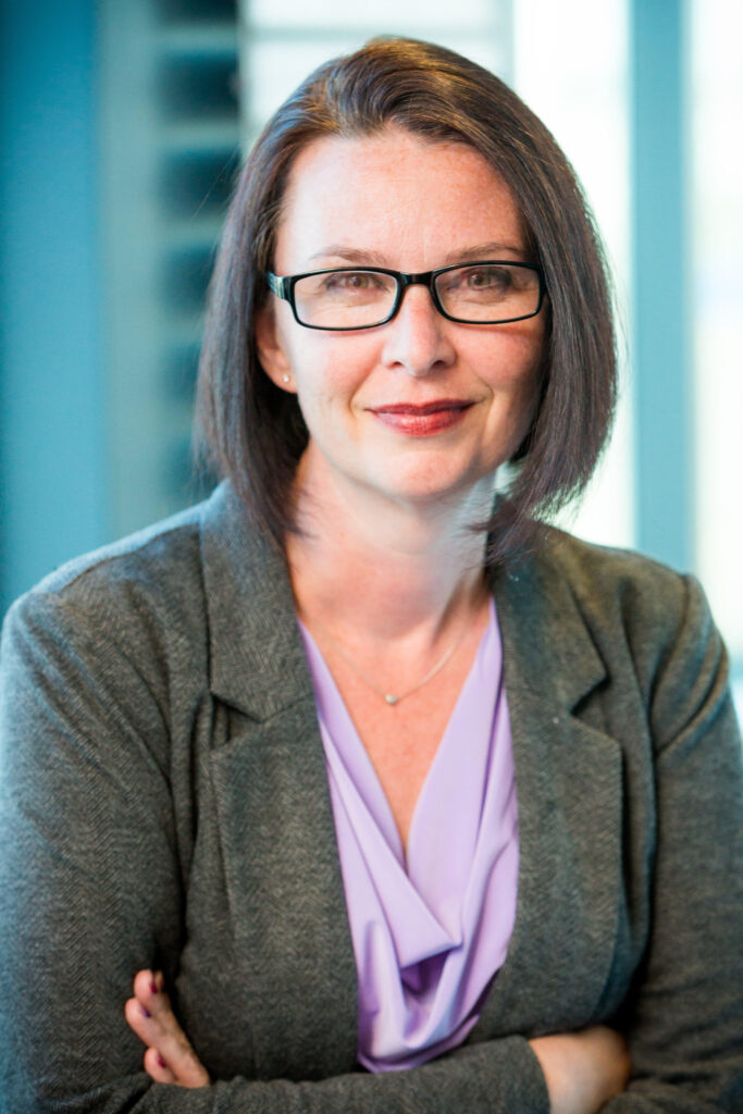 Jennifer White wearing a purple blouse, grey blazer, and black glasses with arms crossed with blue in the background