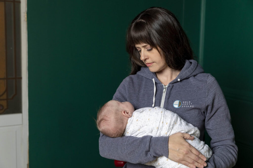 Kay Vorce wearing a heather blue Gold Coast Doulas zip-up hoodie holding a swaddled baby with a dark green wall behind her