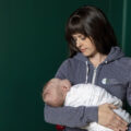 Kay Vorce wearing a heather blue Gold Coast Doulas zip-up hoodie holding a swaddled baby with a dark green wall behind her