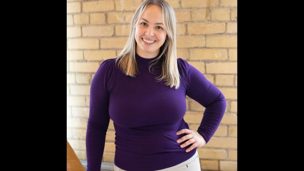 Kristin Mallon poses in front of a brick wall wearing a purple long sleeve top