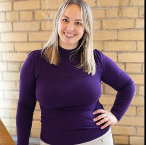 Kristin Mallon poses in front of a brick wall wearing a purple long sleeve top