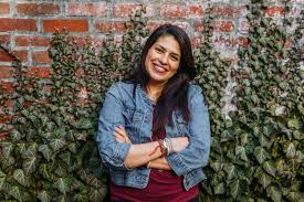 Daniela Procopio of SOLMA Tea poses with her arms crossed in front of a brick wall with greenery wearing a maroon shirt and jean jacket