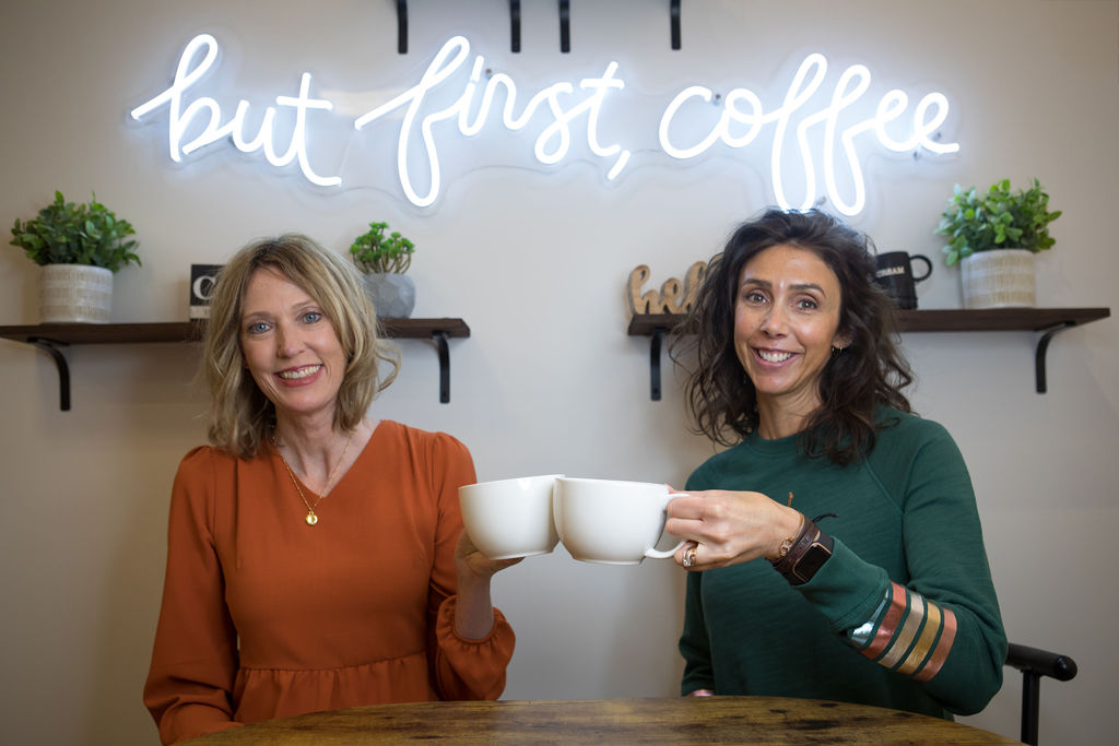 Kristin & Alyssa from Gold Coast Doulas cheers two coffee mugs in front of a neon sign that says "but first, coffee"