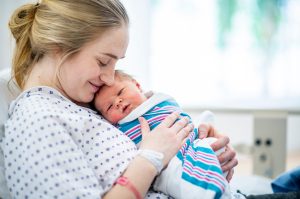 Woman in hospital gown smiling while she holds her newborn baby in a hospital swaddle