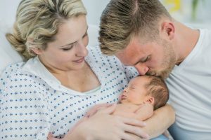 Woman in hospital gown holding her newborn baby while her partner kisses the baby on the forehead