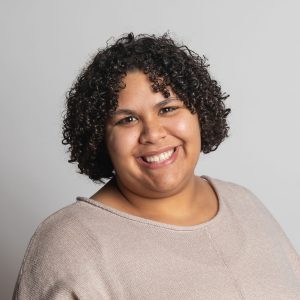 Endira smiling headshot wearing a tan shirt in front of a grey background
