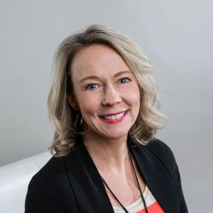 Kelly Emery smiling for a photo wearing a black sweater in front of a grey background