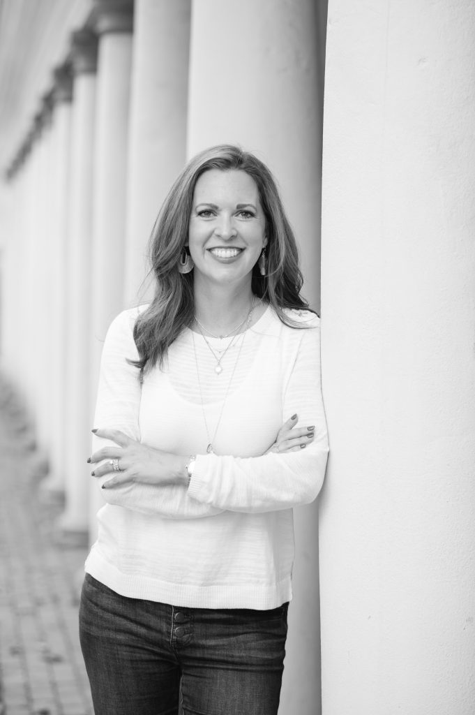 Smiling woman leaning against a column outdoors, arms crossed, wearing a white sweater and jeans.