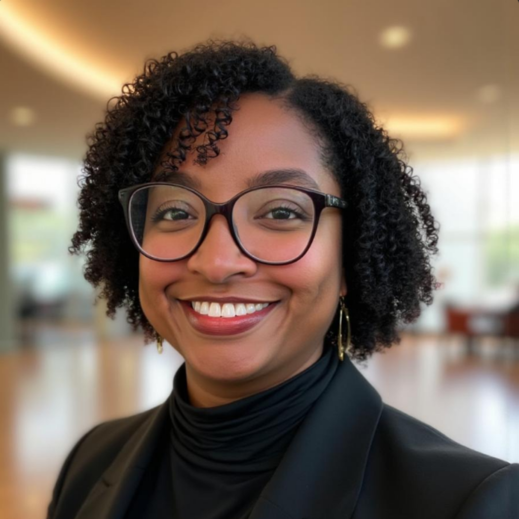 Portrait of a smiling woman with short curly hair, wearing glasses and a black blazer in a bright indoor space, looking at the camera.