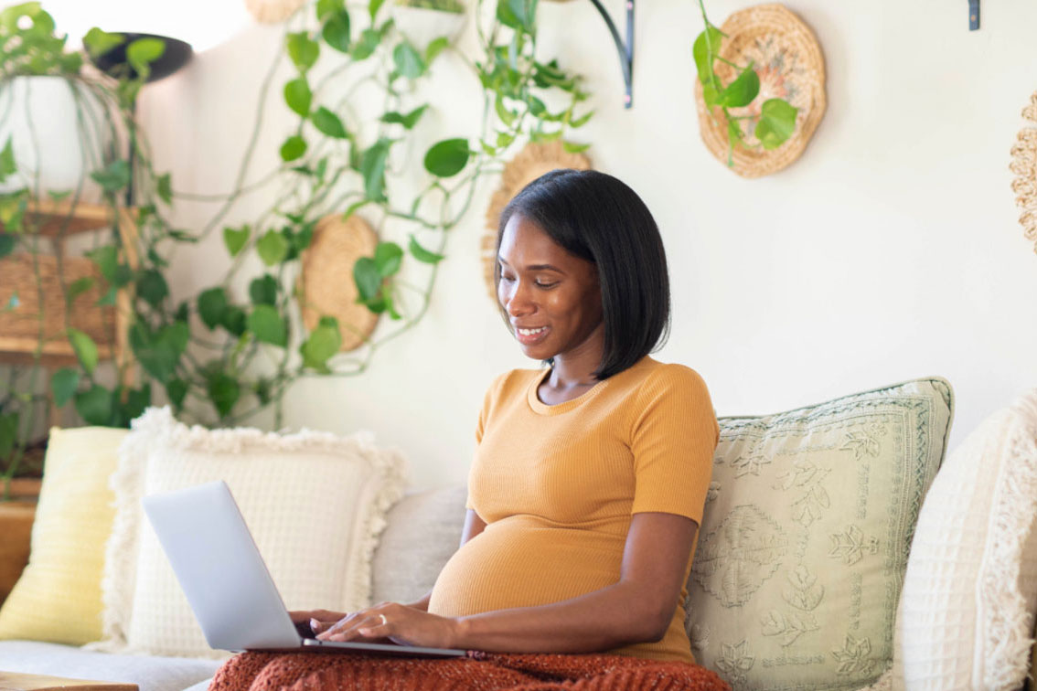 Pregnant woman sitting on a sofa with a laptop, smiling as she types in a plant-filled living room.