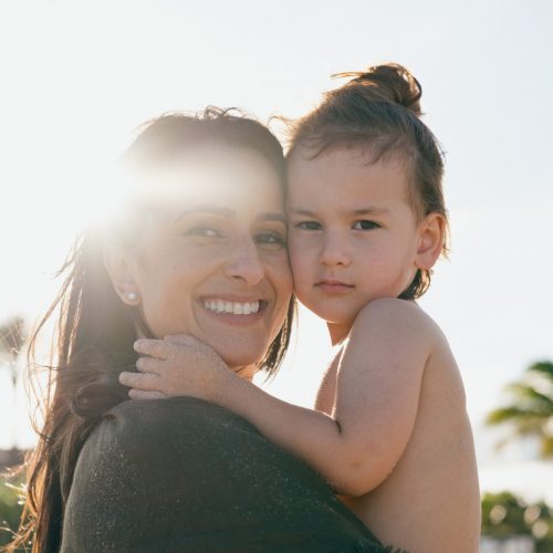 Mother smiles while holding her young daughter at the sunny beach, sun glare in the background.