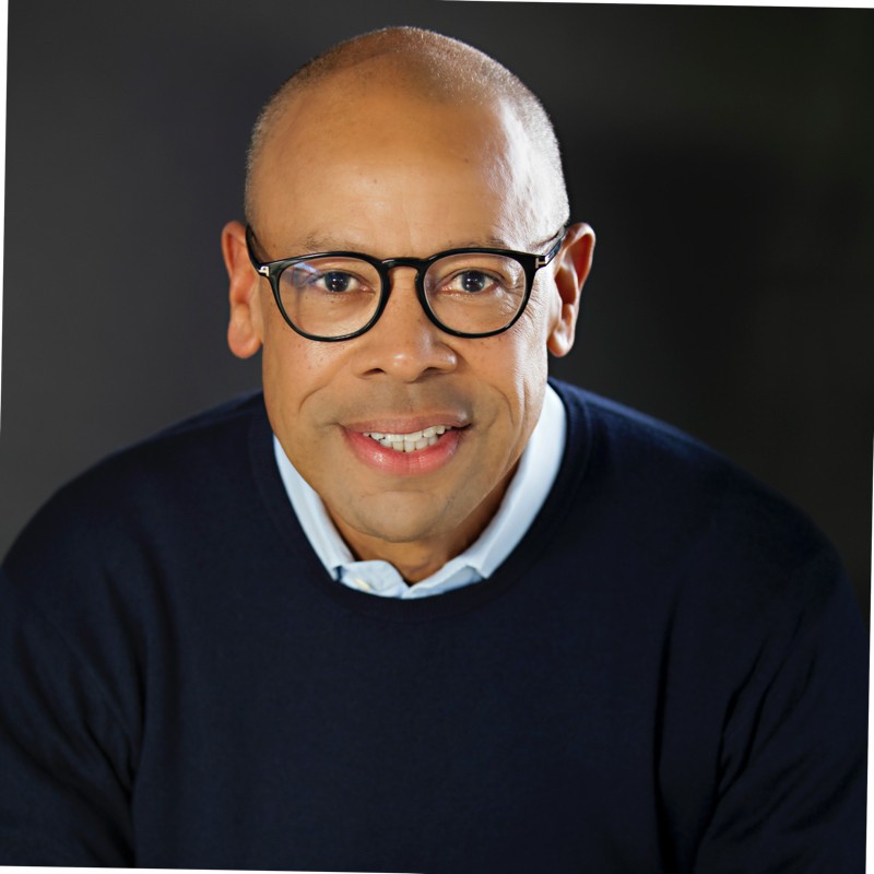Portrait of a smiling man with round glasses, wearing a navy sweater and light blue shirt, against a dark background.
